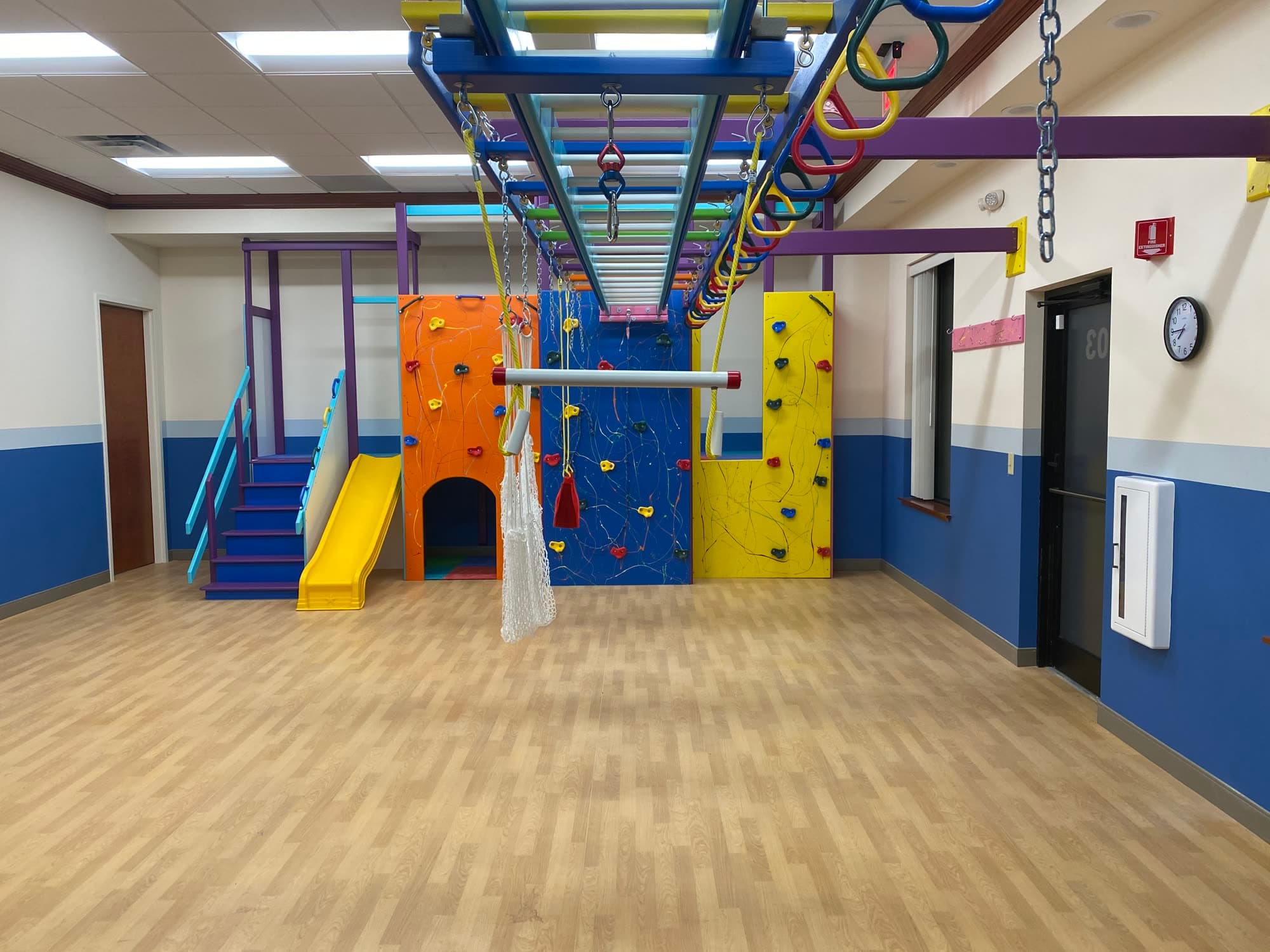 Multi-station sensory gym with orange and yellow climbing walls, yellow slide, and overhead monkey bars