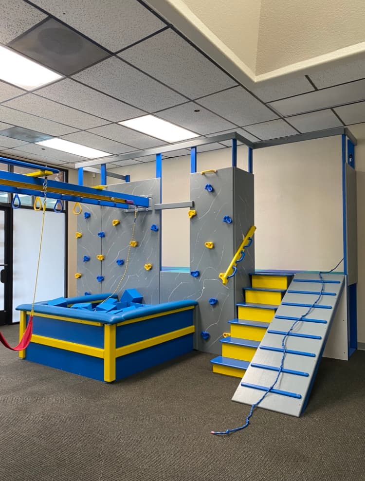 Modern gray clinic sensory gym with climbing wall, blue and yellow ball pit, and ramp ladder