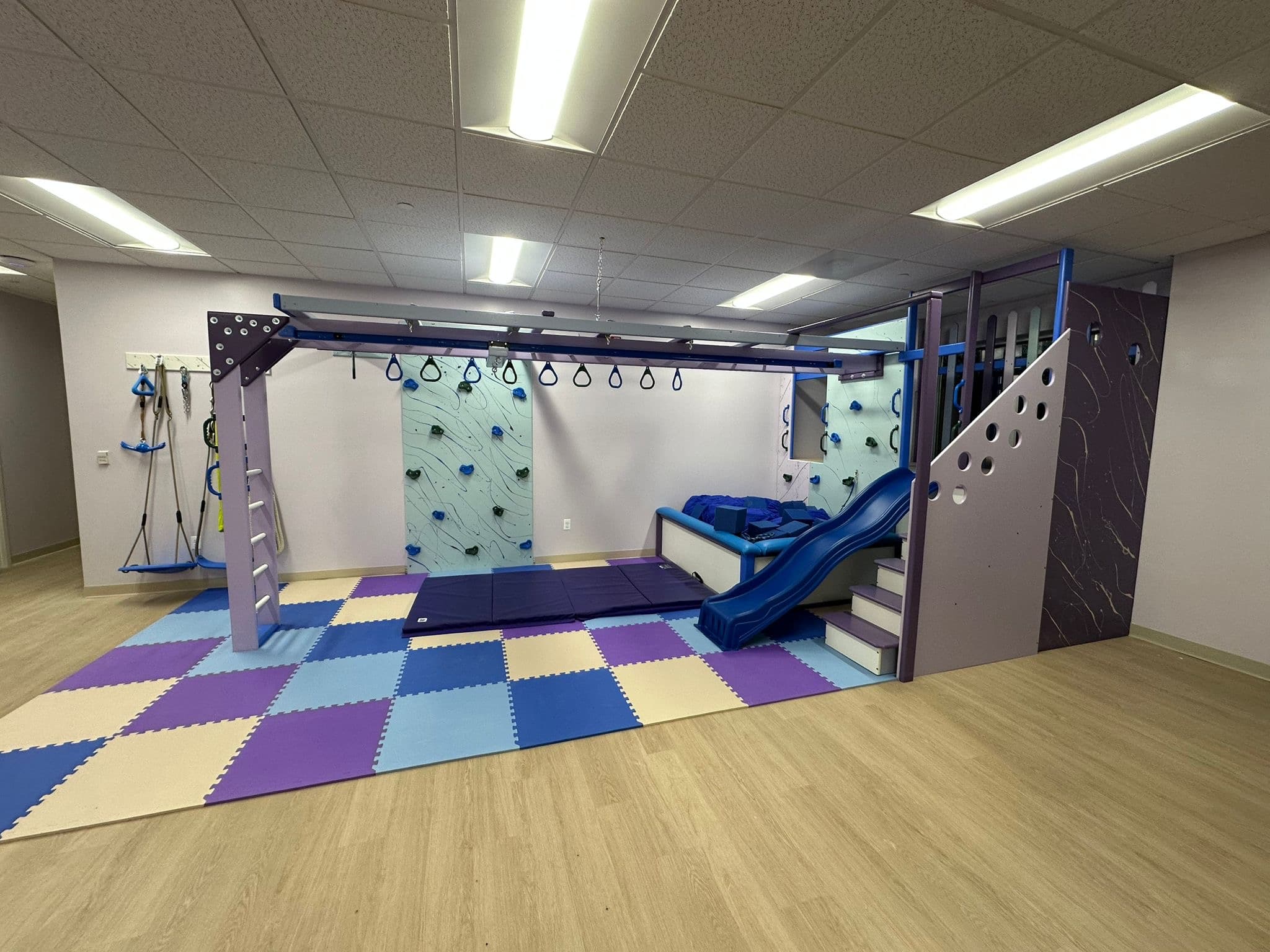 Commercial sensory gym with purple overhead track, rock wall, blue slide, and purple checkered foam floor