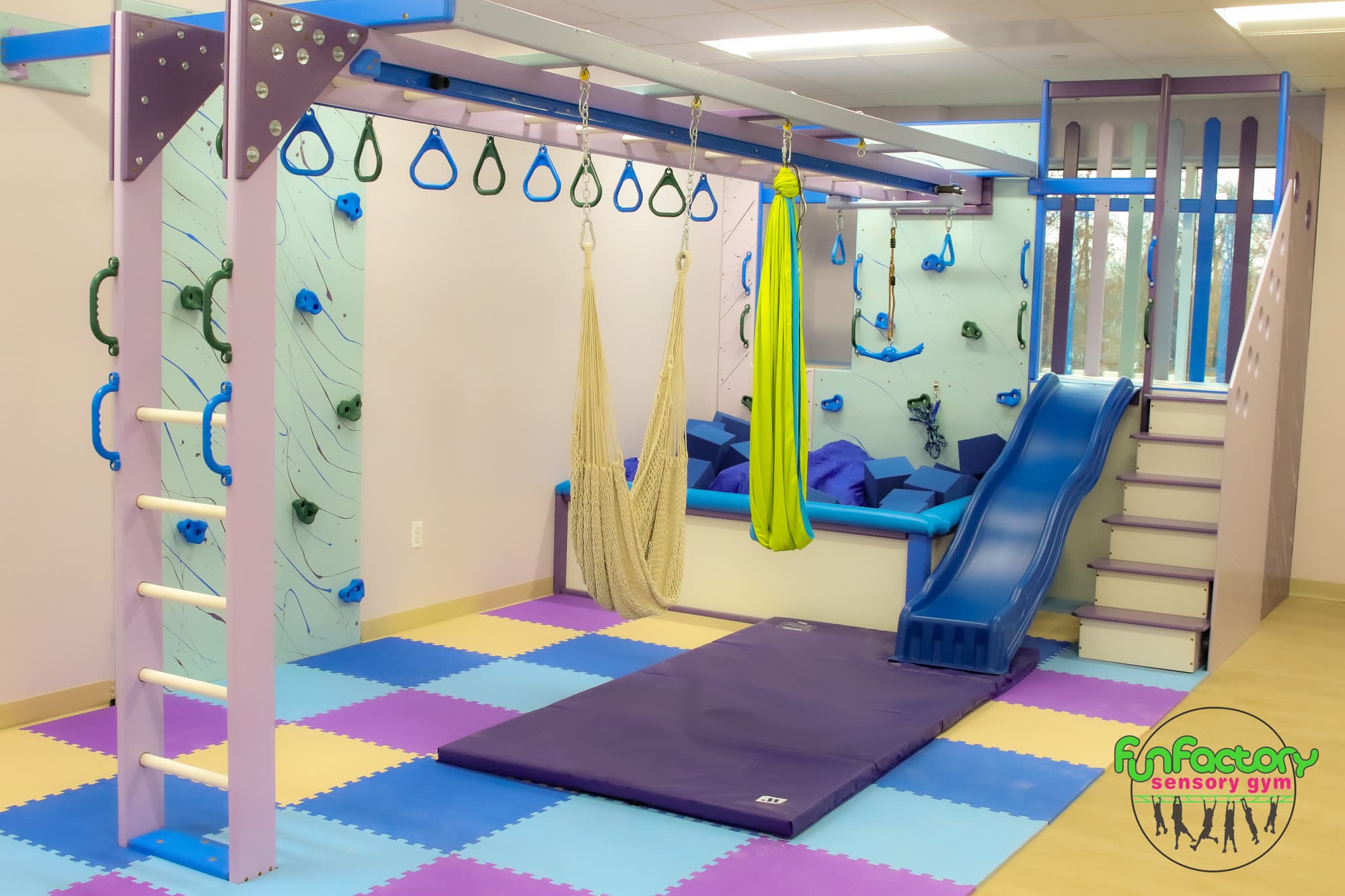 Therapy clinic sensory gym with purple overhead track, lycra swing, blue slide, and climbing wall