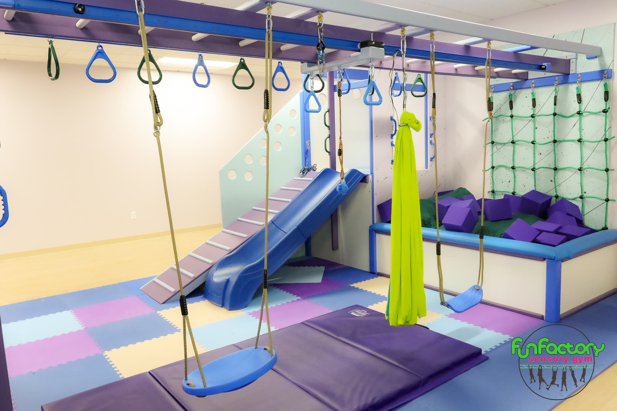 Therapy area with platform swing, green lycra swing, trapeze rings, blue slide, and ball pit