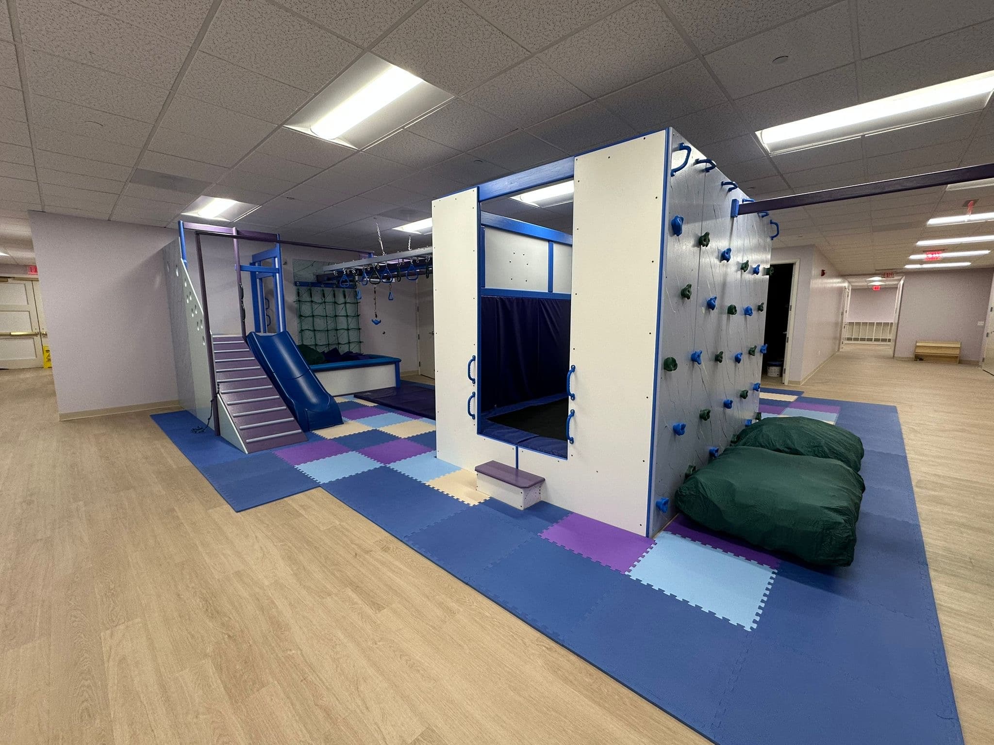 Wide view of a Fun Factory commercial sensory gym with white climbing walls, blue slide, and crash pillows