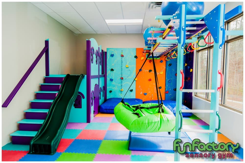 Branded Fun Factory sensory gym with purple stairs, green slide, climbing wall, and green pod swing