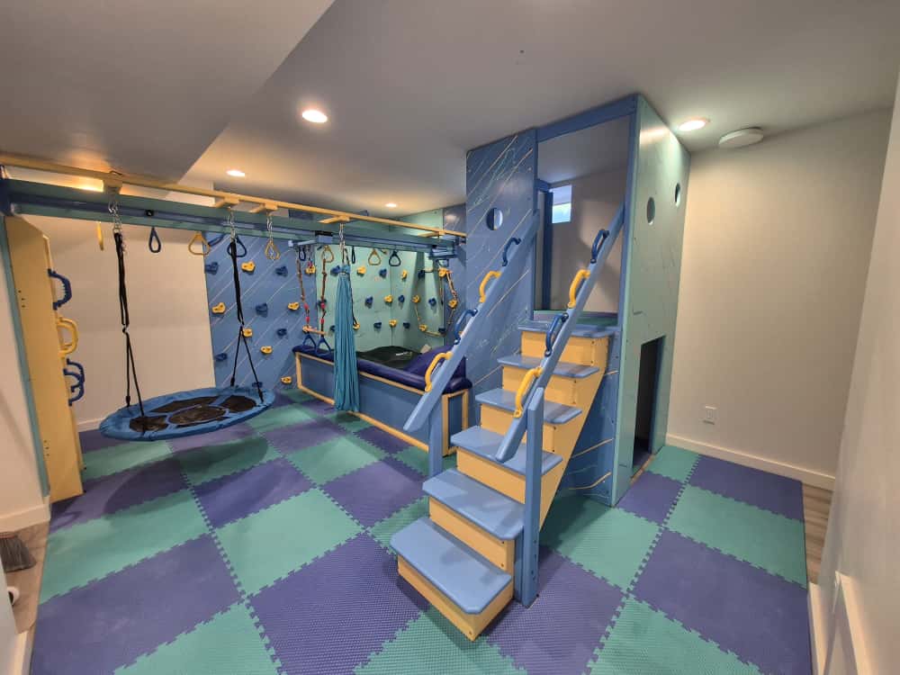 Basement sensory gym install with round platform swing, blue climbing wall, and teal and purple foam floor
