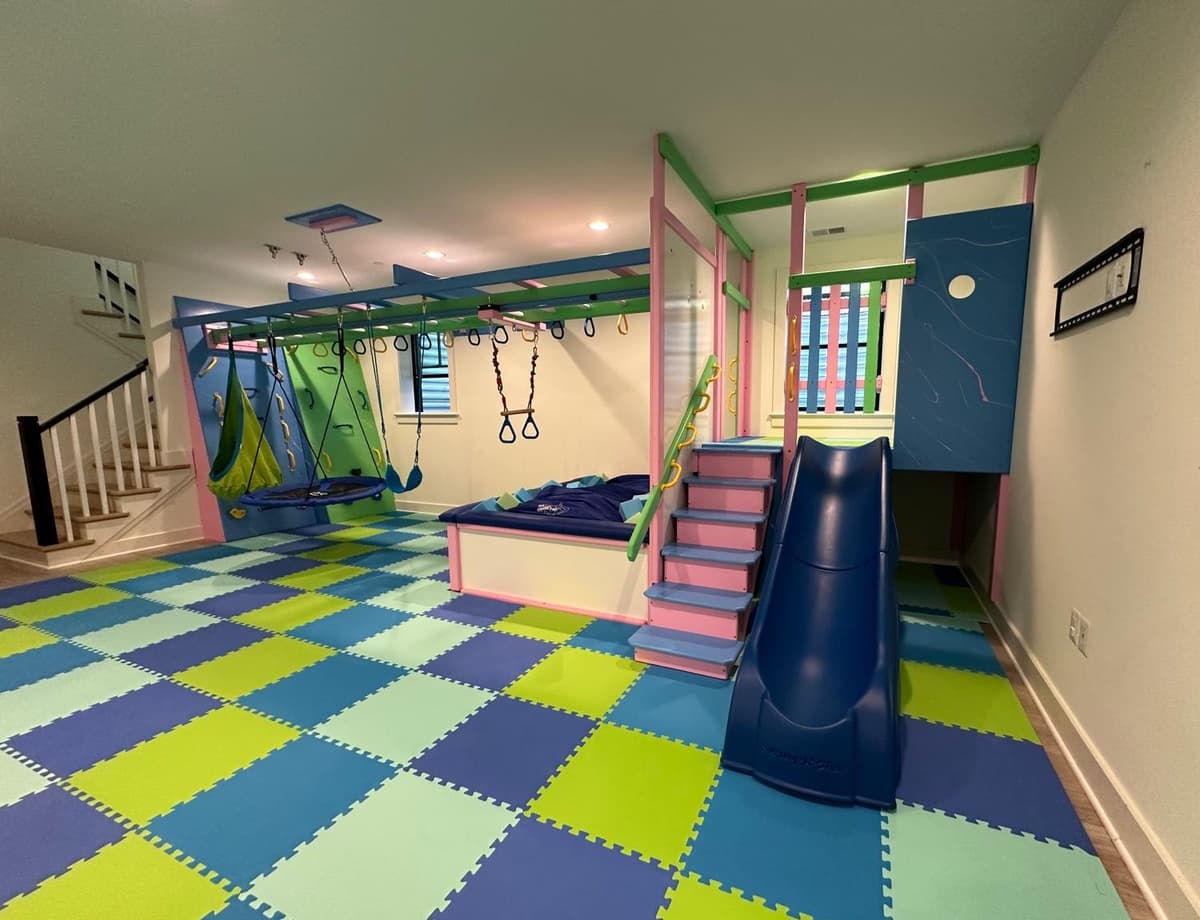 After: same basement with a Fun Factory sensory gym, green checkered foam floor, blue slide, and swings