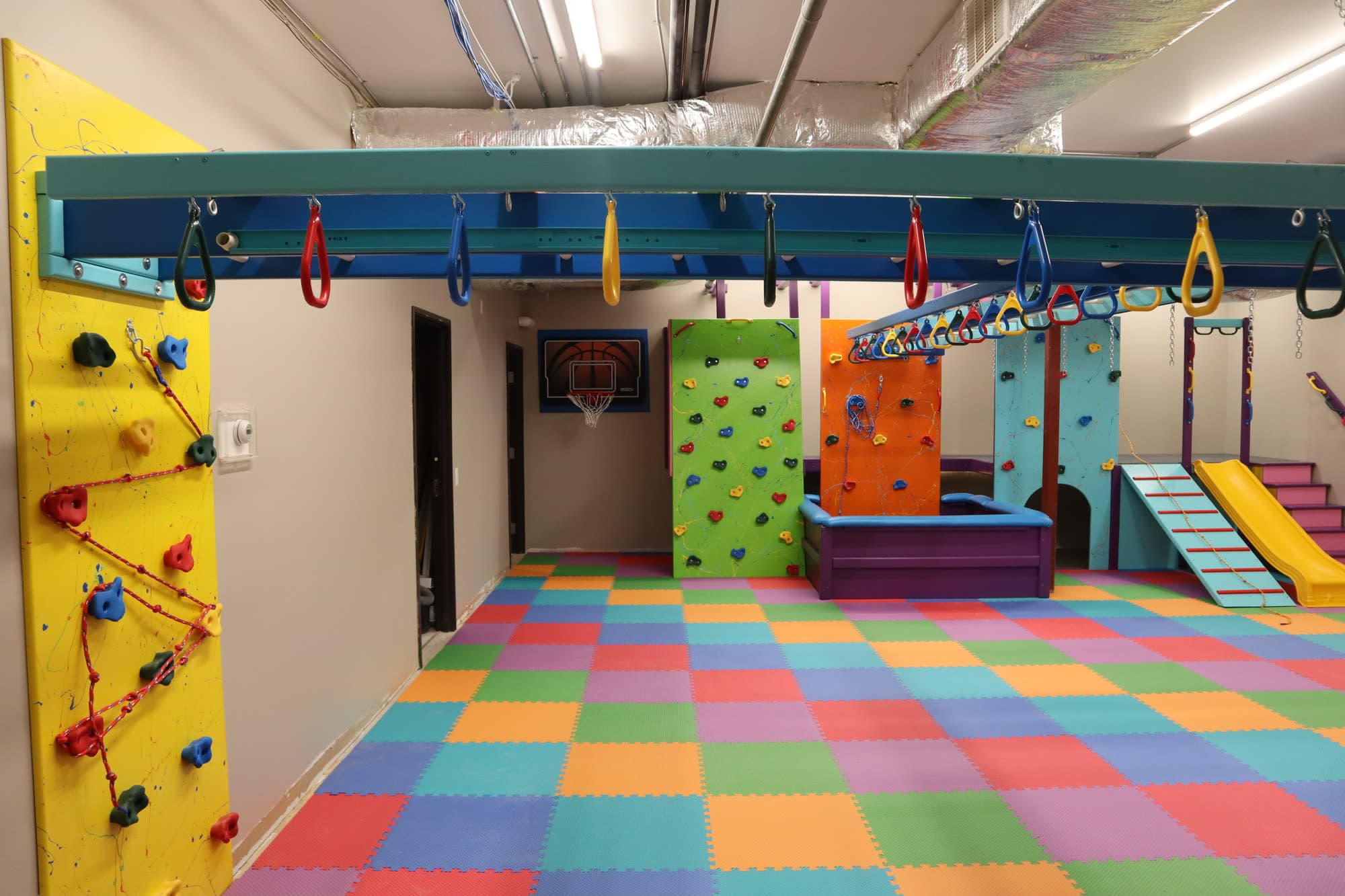 Colorful home sensory gym with yellow climbing wall, overhead trapeze rings, and rainbow foam floor tiles