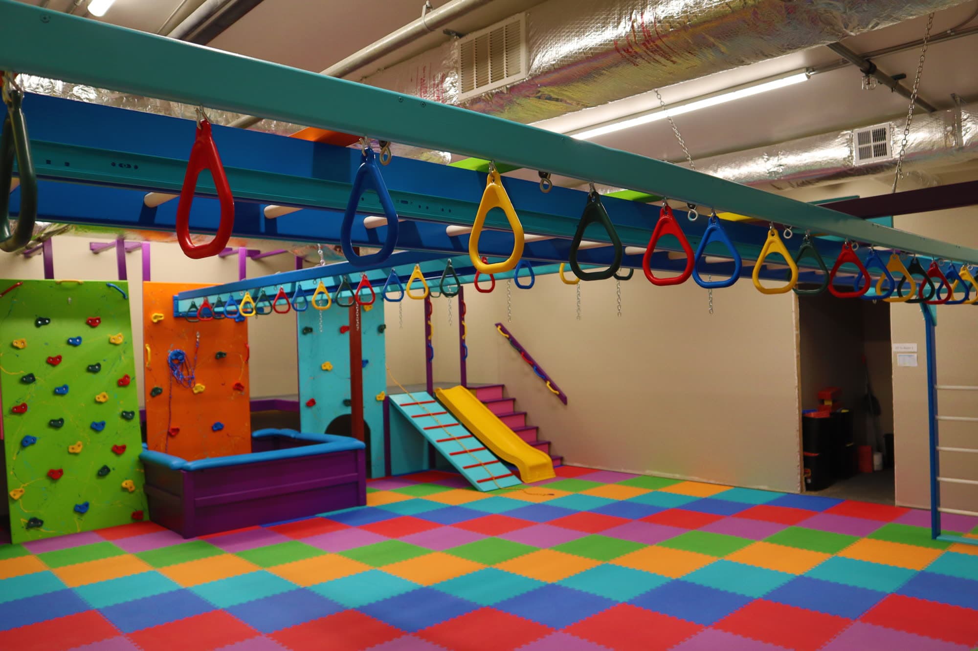 Basement sensory gym with trapeze rings on an overhead track, climbing walls, and rainbow foam floor