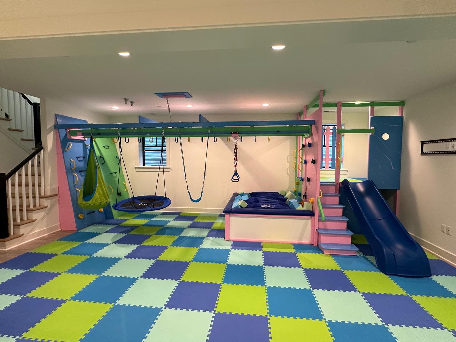 Fun Factory custom home sensory gym in a basement with pod swing, platform swing, blue slide, and green checkered floor