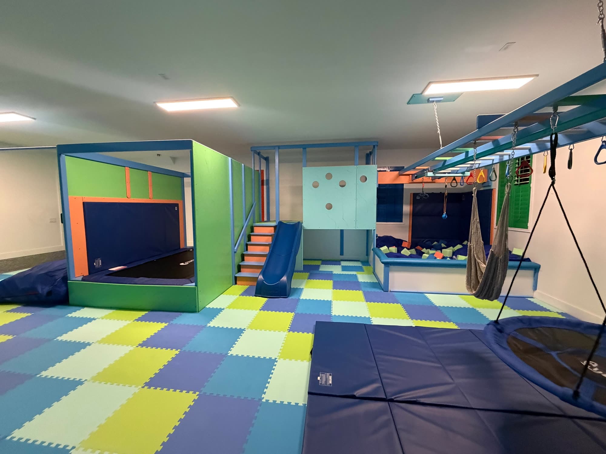 Home playroom sensory gym with blue slide, hammock swing, ball pit, and blue crash mats