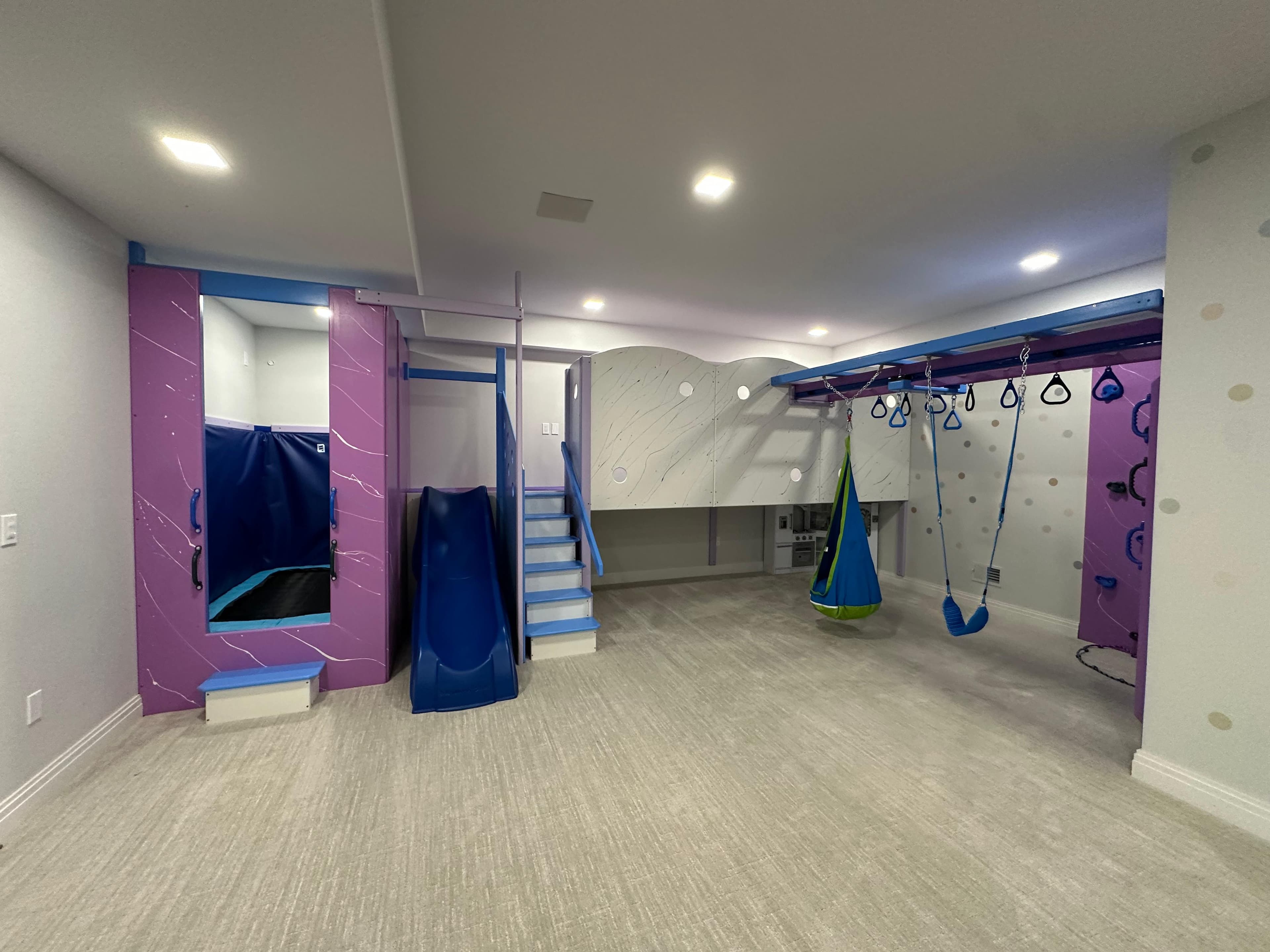 After: same basement with purple climbing wall, blue slide, and pod swing installed in one day by Fun Factory