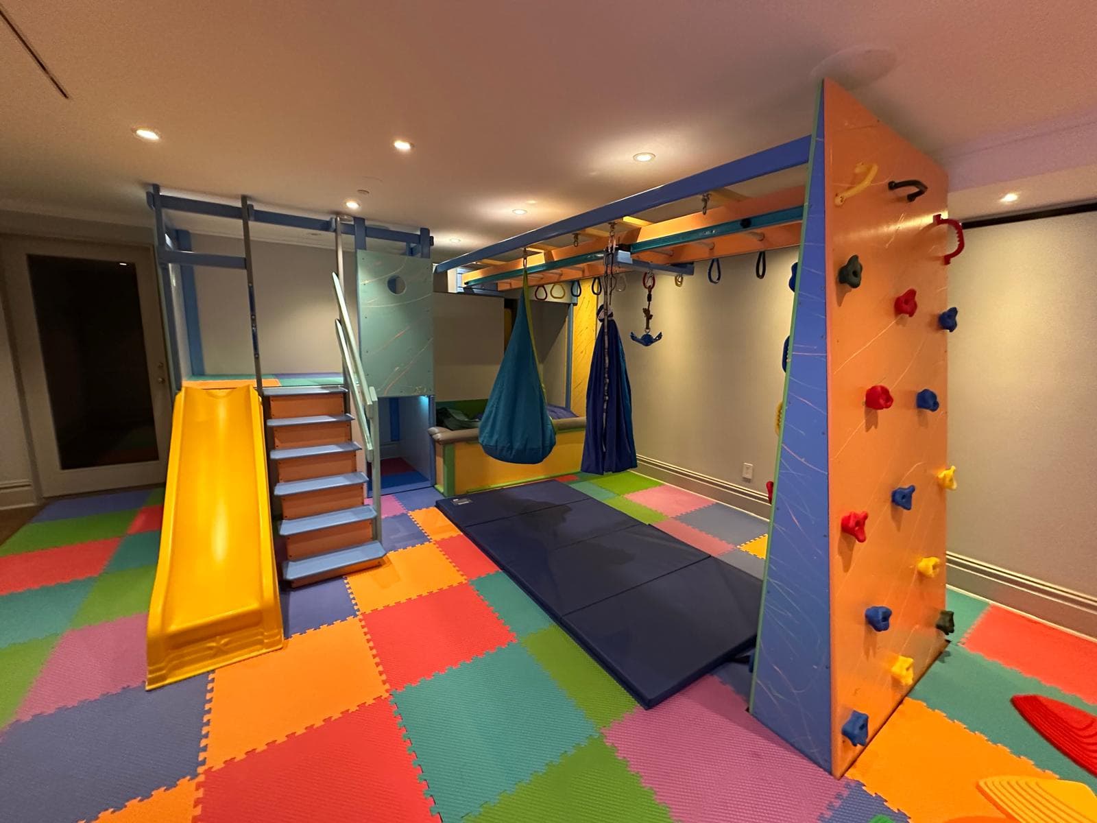 Residential sensory gym with orange climbing wall, yellow slide, hanging pod swing, and rainbow foam floor