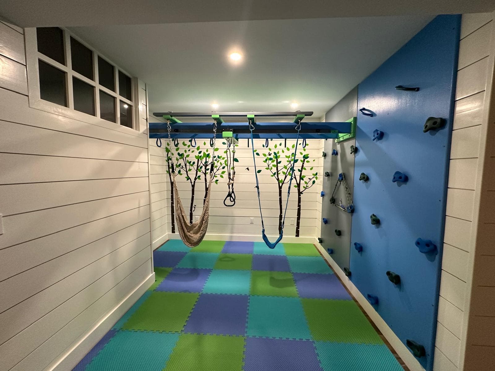 Home therapy gym with hammock swing, blue climbing wall, and tree wall art on white shiplap walls
