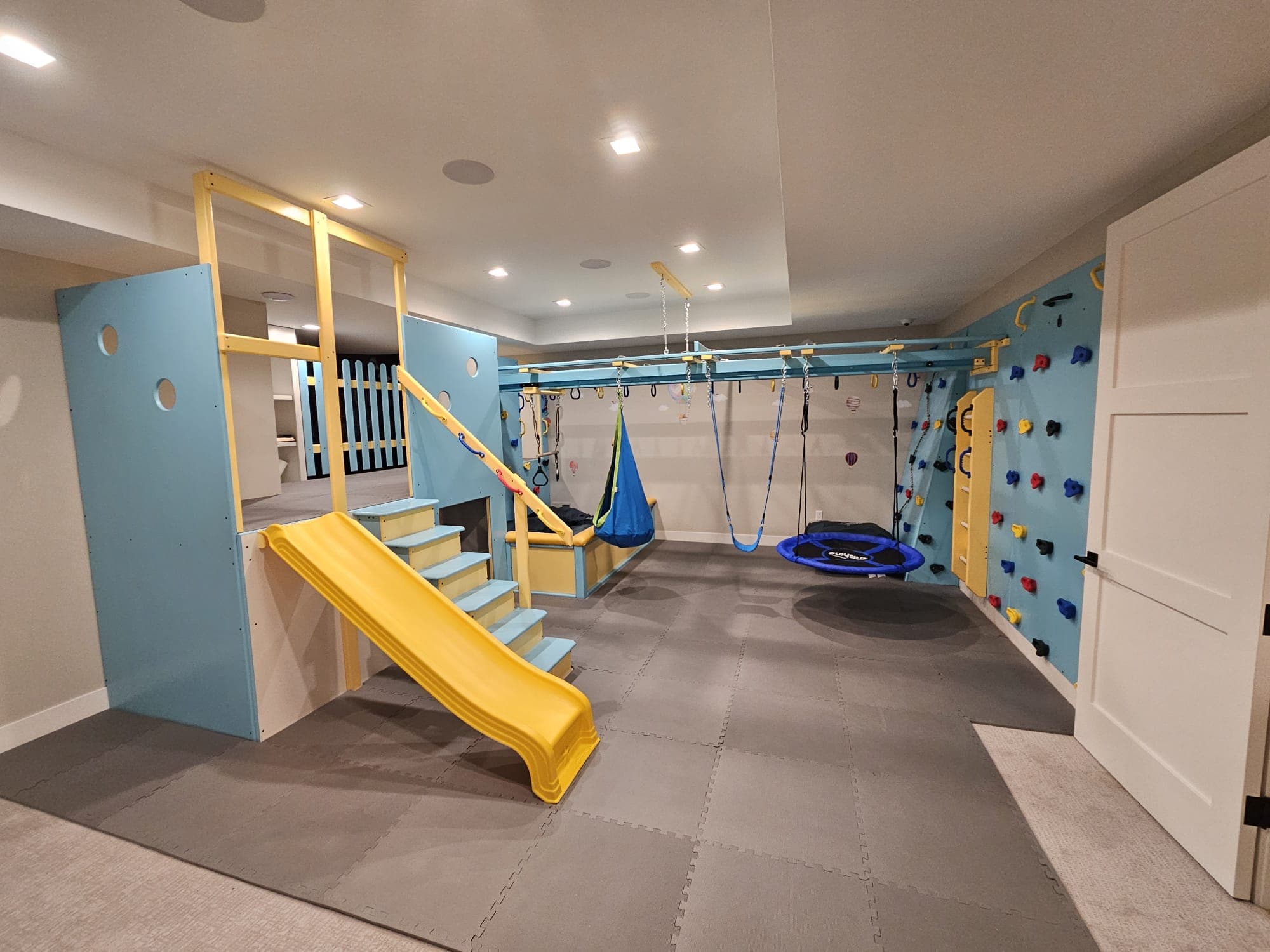 Completed home sensory gym with yellow slide, pod swing, platform swing, and blue climbing wall
