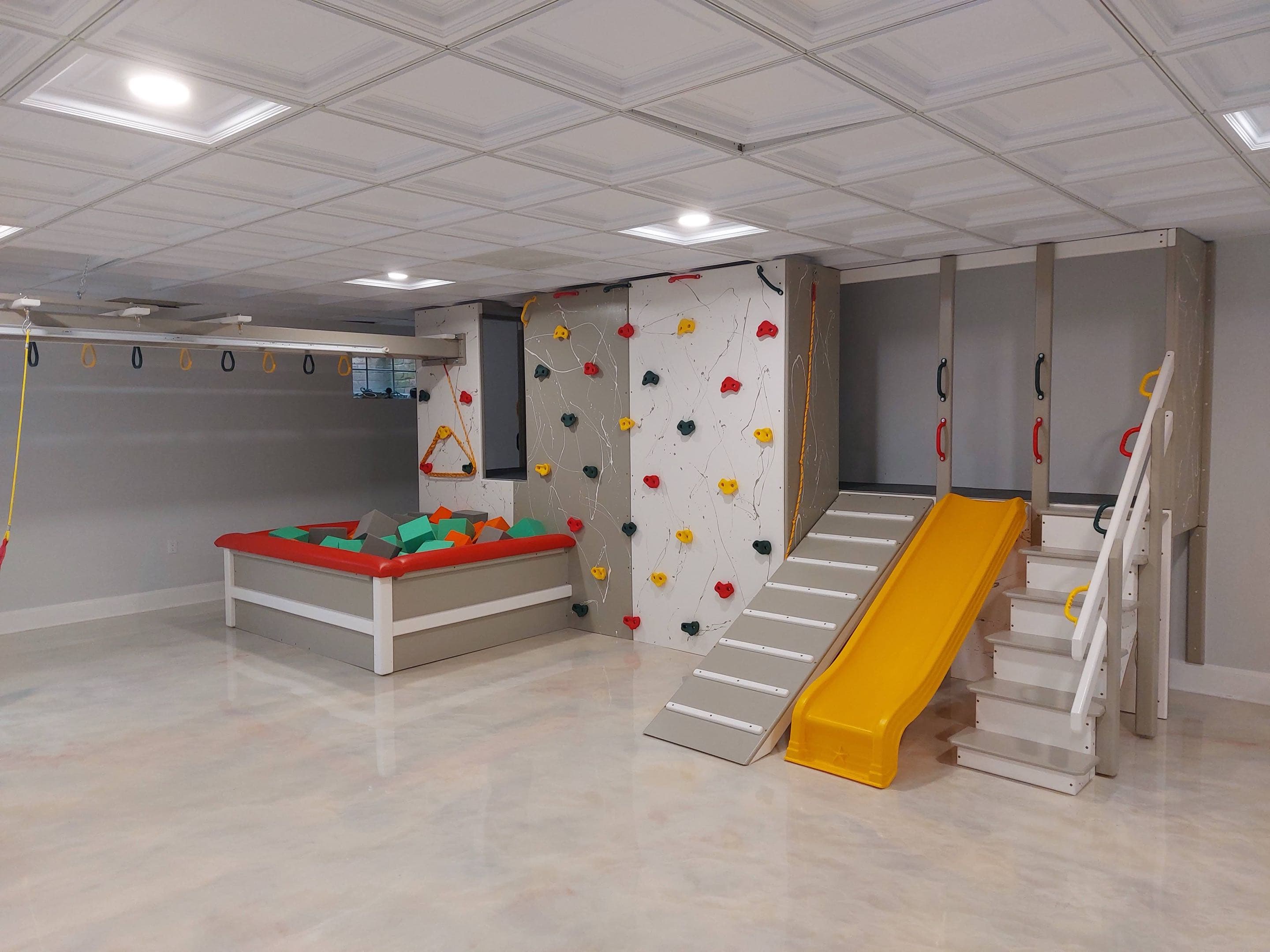 Home basement climbing wall with colorful holds, yellow slide, and foam ball pit by Fun Factory