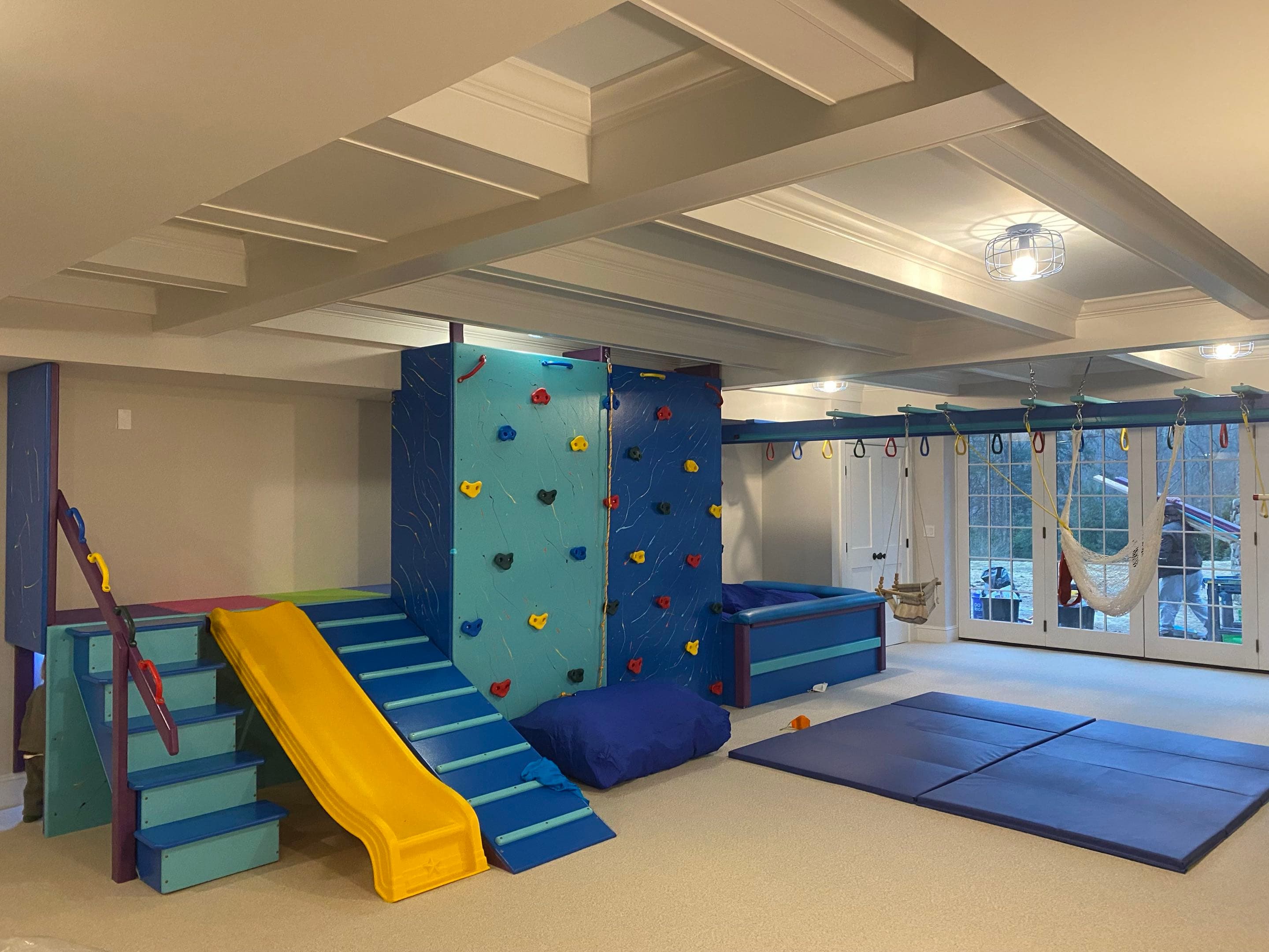 Home play area sensory gym with navy climbing wall, yellow slide, blue crash mats, and hammock swing