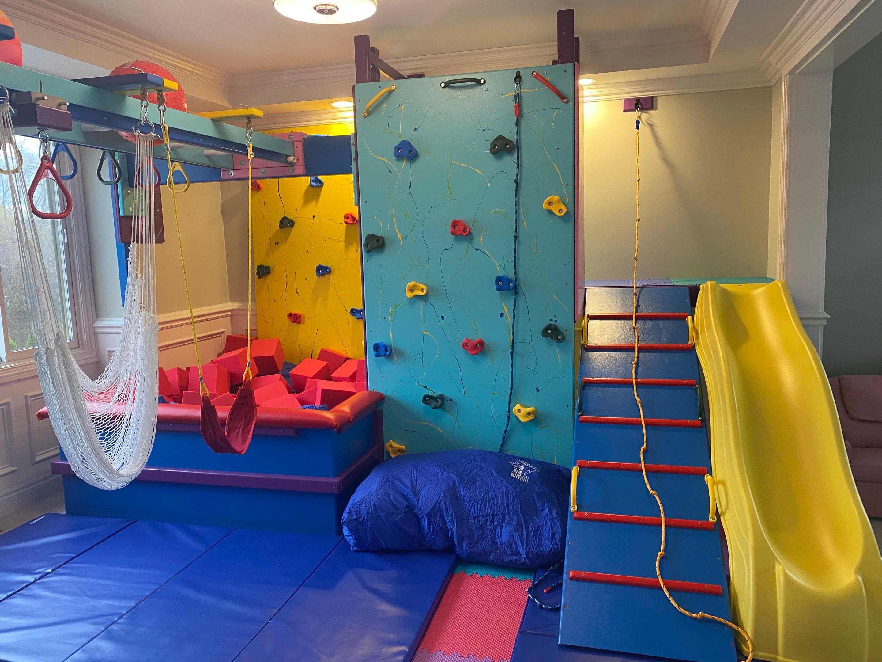 Home sensory gym swing installation with net hammock, teal climbing wall, rope ladder, and yellow slide